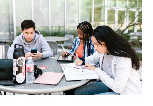 Three students working on school work at an outdoor picnic table on a college campus.