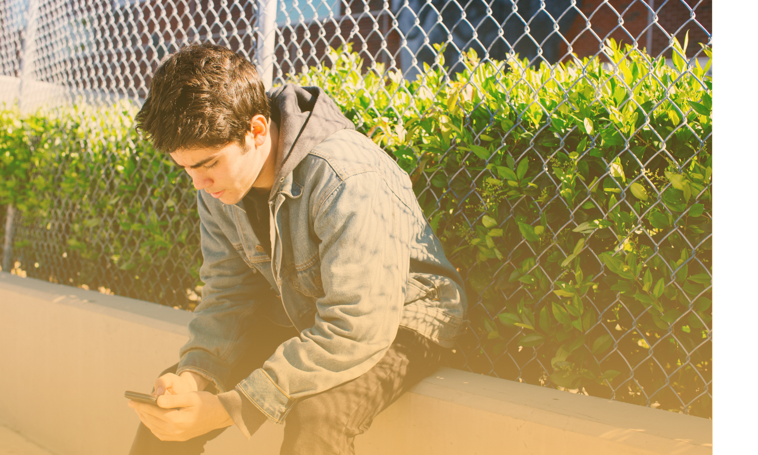 Teen boy sitting by fence outside looking at cell phone