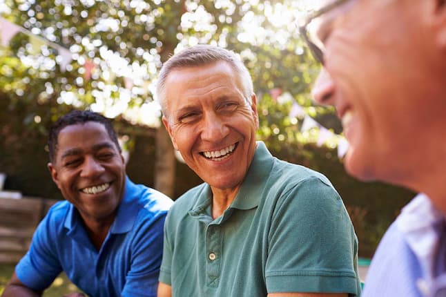 A group of three older men sitting outside and laughing together, enjoying a lighthearted moment in a casual outdoor setting.