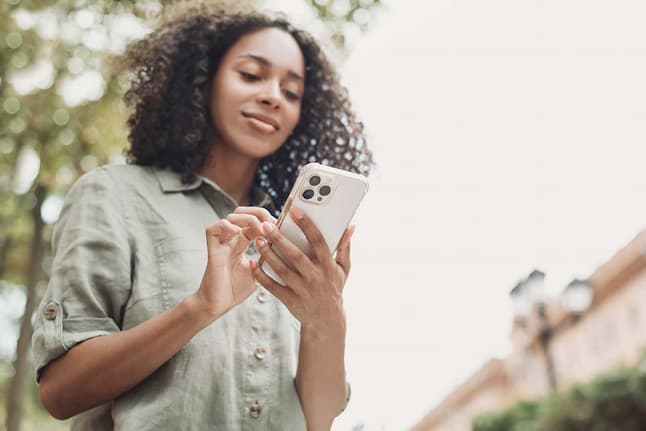 A younger woman standing outdoors, focused on her phone while texting, with a relaxed and content expression.