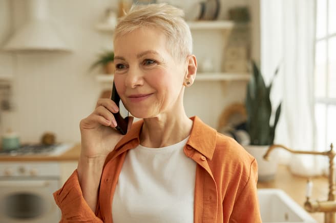 An older woman sitting inside her home, smiling as she talks on the phone.